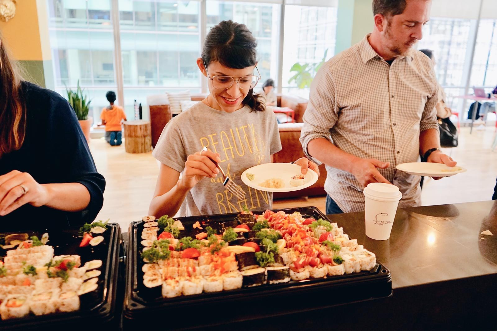 A woman serving herself from sushi trays at an office counter, a colleague behind her.