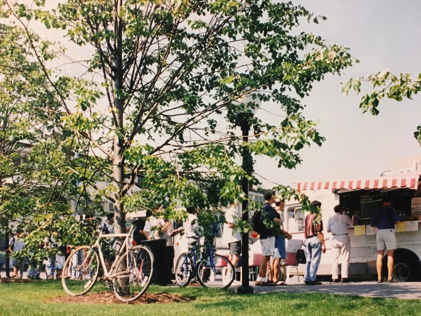 Customers standing at the serving window of the same truck, with a red and white striped awning, bicycles leaning against trees nearby.